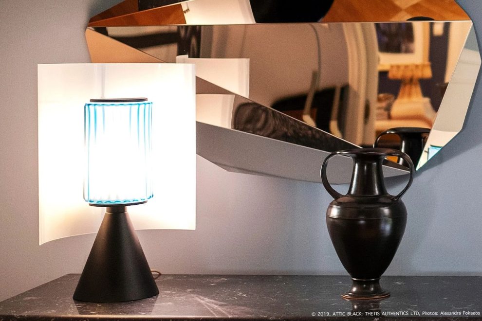 A black amphora resting on a dark tabletop beside a modern lamp with a reflective mirror in the background.
