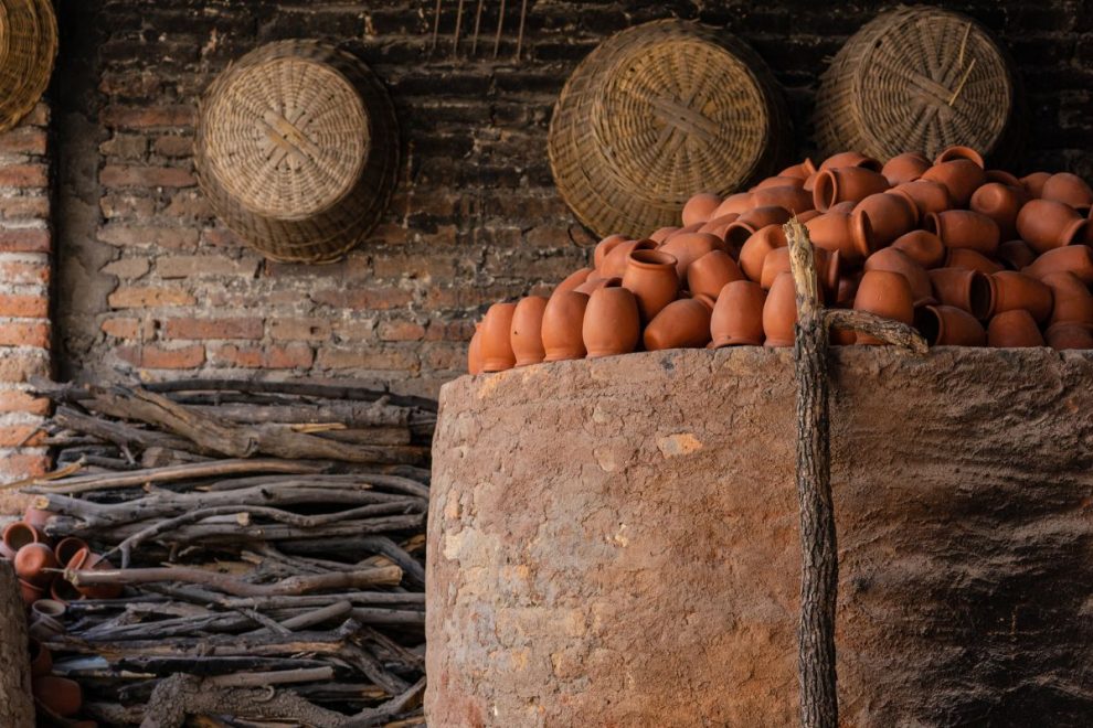 Clay pots stacked near a traditional ancient pottery kiln with firewood stored nearby. 