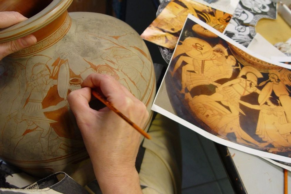 Artist restoring ancient Greek pottery with red-figure battle scene using reference image.